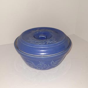 以下が含まれることがあります： 蓋付きの青いセラミックキャセロール皿。 皿は丸い形で、小さなハンドルが付いた蓋が付いています。 皿と蓋はブドウの木の模様で装飾されています。 皿は白い表面に置かれています。
