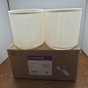 May include: Two cylindrical off-white fabric lamp shades with a visible woven texture. The shades are positioned on top of a brown cardboard shipping box featuring a purple and white label with the text Lampwell and LAMP SHADE.