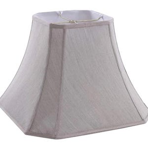 May include: A light gray, square lampshade with a slightly flared shape. The lampshade has a fabric texture and a white interior. The frame is a gold color. The lampshade is designed to fit on a lamp base.