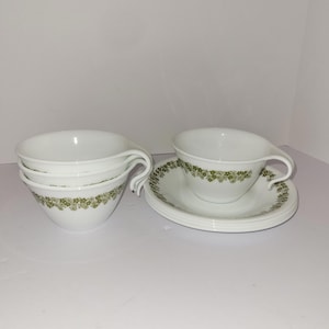 Puede incluir: Tazas y platillos de cerámica blanca con un estampado floral verde. Las tazas tienen un asa curvada y están apiladas en un grupo de tres. Los platillos son redondos y tienen un diseño floral verde a juego alrededor del borde. El juego está sobre una superficie blanca.