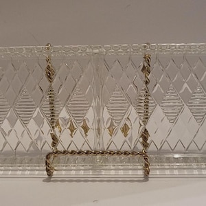 Puede incluir: Fuente de vidrio transparente con un patrón de diamantes y dos secciones. La fuente tiene asas en cada extremo y está sostenida por un soporte de metal dorado. Una pieza decorativa para servir aperitivos.