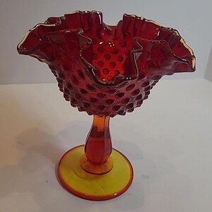 May include: A ruby red hobnail glass pedestal bowl with a ruffled rim and a yellow base. The bowl has a textured surface and a flared, scalloped edge. The stem is a deep orange color.