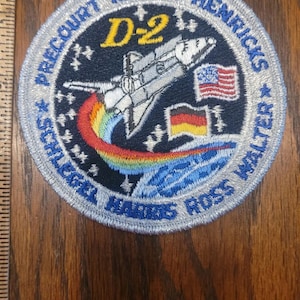 May include: Embroidered patch featuring a space shuttle, rainbow, and flags of the United States and Germany. The patch is circular with text around the edge, including names and "D-2". The background is dark blue.