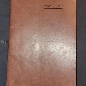 May include: A vintage, brown leather-bound book with the text "REVISED 1912" printed in black. The book shows signs of wear, with creases and a slightly rounded corner. The cover is a solid, warm brown colour.
