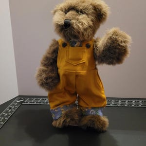 May include: A brown teddy bear wearing yellow overalls with a pocket. The overalls have blue floral patterned cuffs and a matching patch on the chest. The bear has a black nose and is waving its right paw.