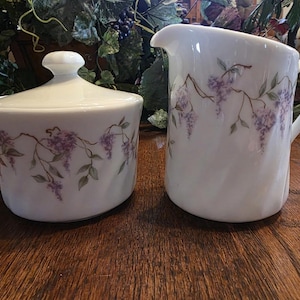 May include: White ceramic sugar bowl with lid and a creamer pitcher. Both pieces are decorated with a purple wisteria floral design. The sugar bowl is round with a small handle on the lid. The creamer has a handle and a spout.