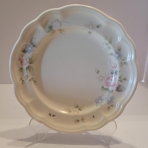 Puede incluir: Un plato de cerámica blanco con borde festoneado y un diseño floral con flores rosas, azules y verdes.