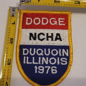 May include: A vintage patch with a shield shape, featuring the text "DODGE NCHA DUQUOIN ILLINOIS 1976." The patch has a red, white, and blue color scheme with a gold border. The patch measures approximately 3 inches in length.