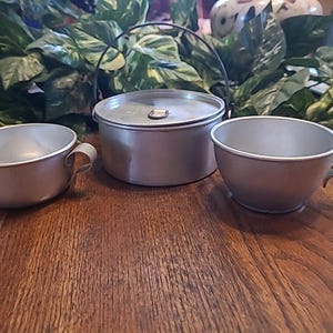 May include: A set of vintage aluminum camping cookware, including a small pot with a handle, and two cups with handles. The items are silver-colored and sit on a wooden surface, with green foliage in the background.