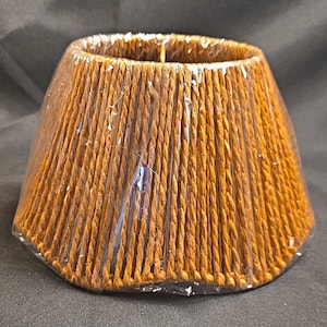 May include: A conical lampshade constructed from vertical rows of twisted brown cord. The lampshade is wrapped in a clear, protective plastic film that covers the entire exterior surface, maintaining its shape and structure.