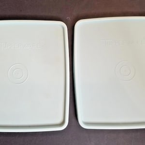 Puede incluir: Dos tapas cuadradas Tupperware de color azul claro. Cada tapa tiene esquinas redondeadas, un borde ligeramente elevado y un diseño circular en el centro. La palabra "Tupperware" está impresa en cada tapa.