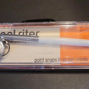 Puede incluir: Un bolígrafo blanco en una caja de plástico transparente con detalles en naranja y blanco. El bolígrafo tiene un clip plateado y un dije plateado. El texto en la caja dice "cool riter" y "point snaps in as pen winds back."