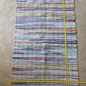 May include: Rectangular woven rug with a colorful striped pattern. The rug is approximately 30 inches long and 20 inches wide, with a fringe on the short ends. The rug features a mix of blue, red, yellow, and pink hues.