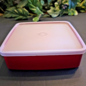 Puede incluir: Un recipiente cuadrado rojo con una tapa rosa claro. La tapa tiene un centro ligeramente elevado con un diseño circular. El recipiente está hecho de plástico y parece ser un artículo para guardar alimentos.