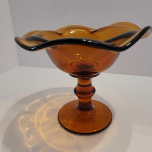 Puede incluir: Cuenco de pedestal de vidrio color ámbar con borde ondulado y ribete negro. El cuenco tiene una base redonda y un tallo decorativo. El cuenco de vidrio es probablemente vintage y adecuado para servir fruta o exhibir objetos decorativos.
