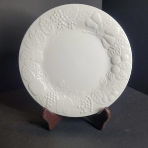 Peut inclure: Une assiette en céramique blanche avec un motif en relief de fruits et de feuilles sur le bord. L'assiette est ronde et repose sur un support marron. Le motif comprend des raisins et des poires.