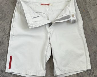 May include: Pale white shorts with a button and zip fastening. The shorts feature a red stripe detail on the side and the brand name "PRADA" is visible on the waistband. The shorts are made of a light-coloured fabric.