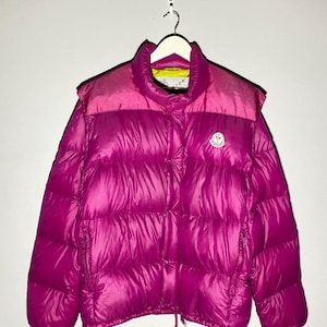 Puede incluir: Una chaqueta acolchada rosa con una cremallera amarilla y un parche de logotipo blanco. La chaqueta tiene un diseño acolchado y un cuello alto.