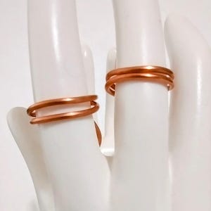 Puede incluir: Dos anillos de color cobre se muestran en una exhibici&oacute;n blanca en forma de mano. Los anillos tienen un dise&ntilde;o simple y minimalista, con una apariencia de doble banda. Los anillos est&aacute;n hechos de un material met&aacute;lico brillante, y la est&eacute;tica general es moderna y elegante.