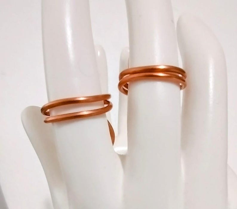 Puede incluir: Dos anillos de color cobre se muestran en una exhibici&oacute;n blanca en forma de mano. Los anillos tienen un dise&ntilde;o simple y minimalista, con una apariencia de doble banda. Los anillos est&aacute;n hechos de un material met&aacute;lico brillante, y la est&eacute;tica general es moderna y elegante.
