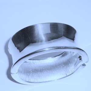 May include: A silver bangle bracelet with a smooth, polished finish on the top and a textured, brushed finish on the bottom.