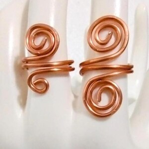 Puede incluir: Dos anillos de alambre de cobre, cada uno con un dise&ntilde;o en espiral. Los anillos se muestran sobre una superficie blanca, mostrando sus intrincados detalles hechos a mano. Los anillos tienen un tono marr&oacute;n rojizo c&aacute;lido.