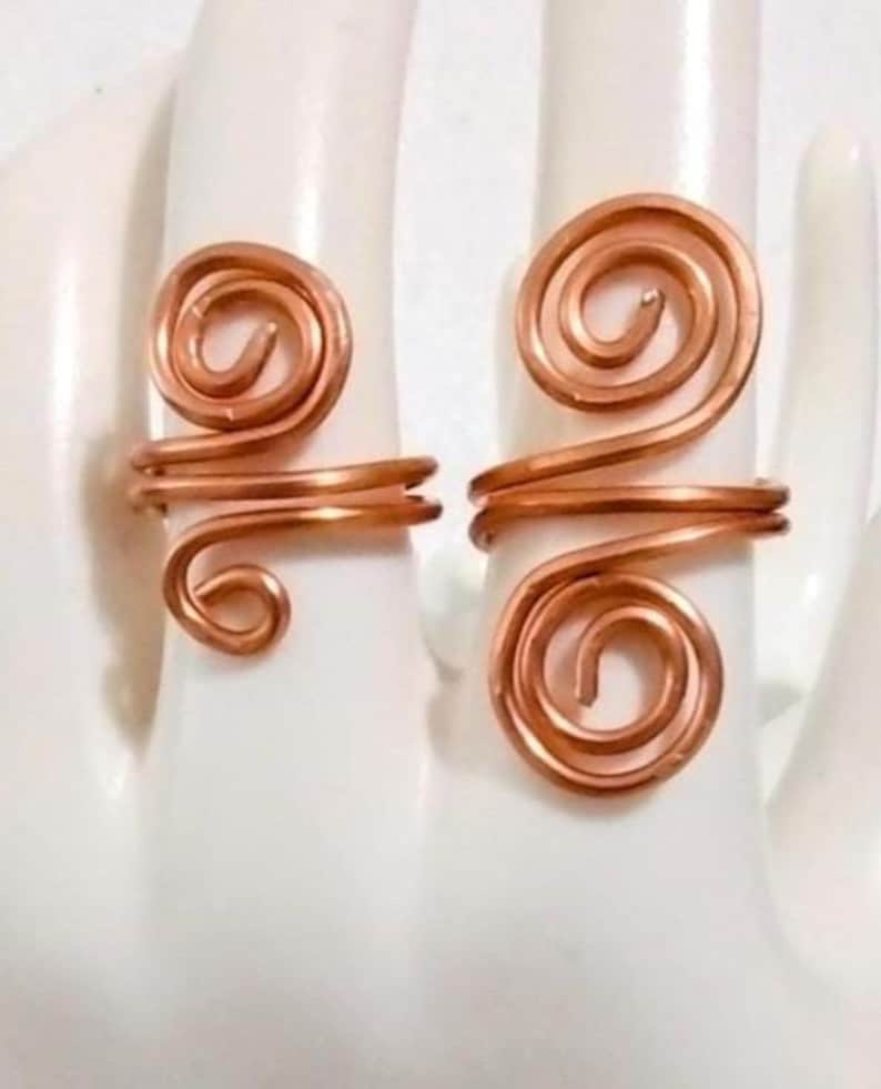 Puede incluir: Dos anillos de alambre de cobre, cada uno con un dise&ntilde;o en espiral. Los anillos se muestran sobre una superficie blanca, mostrando sus intrincados detalles hechos a mano. Los anillos tienen un tono marr&oacute;n rojizo c&aacute;lido.