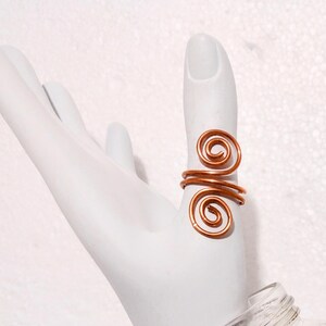 Puede incluir: Un anillo de alambre de color cobre con un dise&ntilde;o en espiral. El anillo est&aacute; envuelto alrededor del dedo, con dos acentos en espiral. El anillo se muestra en una mano blanca.