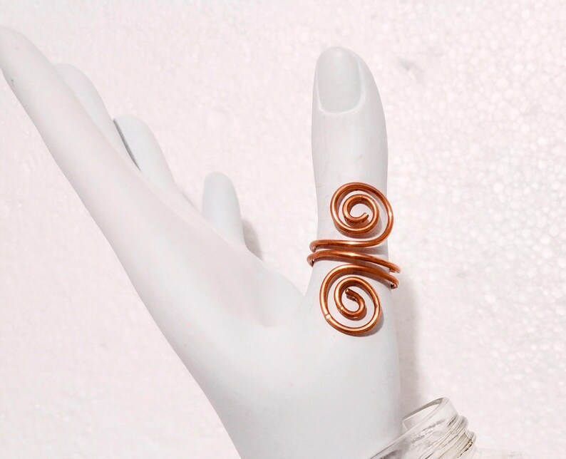Puede incluir: Un anillo de alambre de color cobre con un dise&ntilde;o en espiral. El anillo est&aacute; envuelto alrededor del dedo, con dos acentos en espiral. El anillo se muestra en una mano blanca.