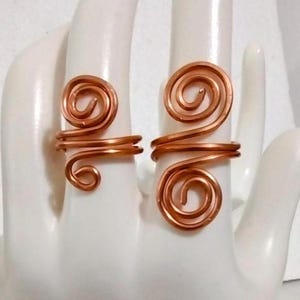 Puede incluir: Dos anillos de alambre de color cobre, cada uno con un dise&ntilde;o en espiral. Los anillos se muestran en una mano blanca, destacando el intrincado trabajo de alambre y la forma &uacute;nica de los anillos. Son joyas hechas a mano.