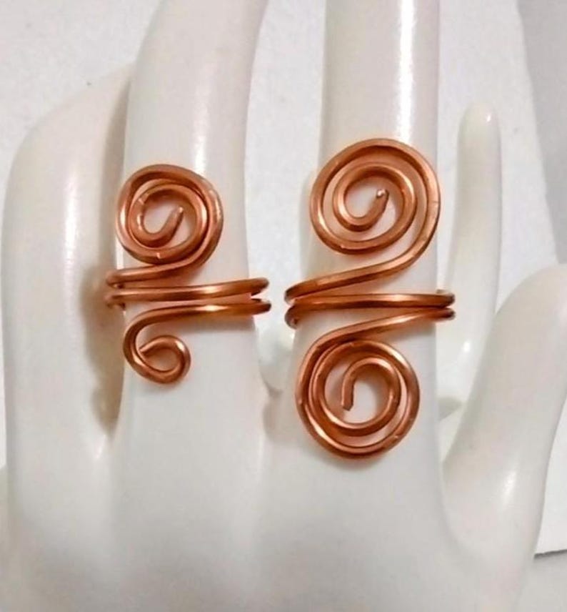 Puede incluir: Dos anillos de alambre de color cobre, cada uno con un dise&ntilde;o en espiral. Los anillos se muestran en una mano blanca, destacando el intrincado trabajo de alambre y la forma &uacute;nica de los anillos. Son joyas hechas a mano.