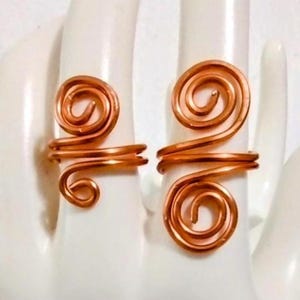 Puede incluir: Dos anillos de alambre color cobre, cada uno con un dise&ntilde;o en espiral. Los anillos se exhiben sobre una superficie blanca, mostrando sus intrincados detalles hechos a mano. El dise&ntilde;o en espiral a&ntilde;ade un toque art&iacute;stico &uacute;nico a la joyer&iacute;a.