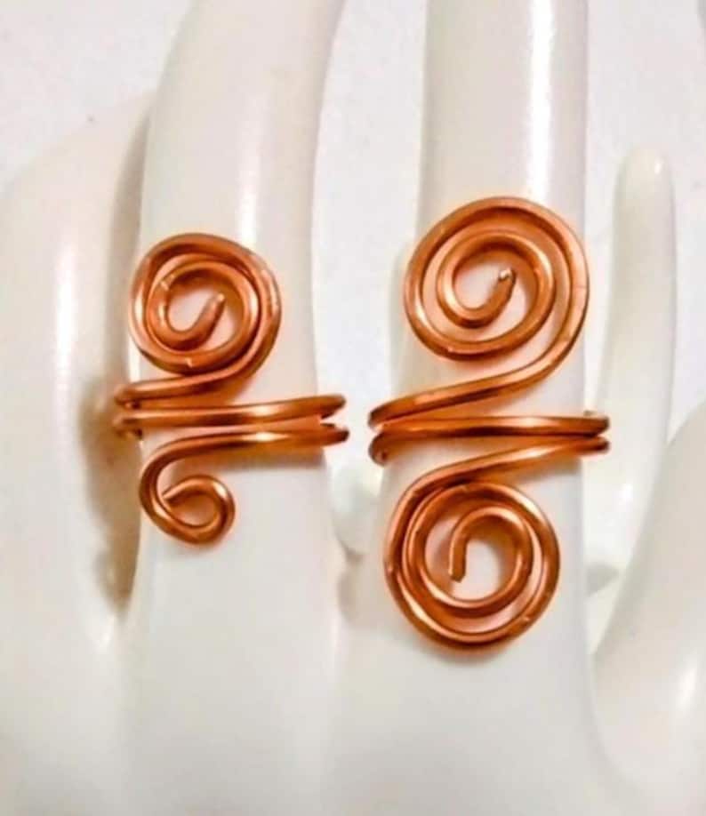 Puede incluir: Dos anillos de alambre color cobre, cada uno con un dise&ntilde;o en espiral. Los anillos se exhiben sobre una superficie blanca, mostrando sus intrincados detalles hechos a mano. El dise&ntilde;o en espiral a&ntilde;ade un toque art&iacute;stico &uacute;nico a la joyer&iacute;a.