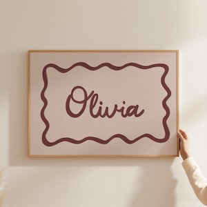 Puede incluir: Una impresión artística enmarcada con el nombre "Olivia" en fuente cursiva. El nombre es de color burdeos oscuro, sobre un fondo rosa claro. La impresión está enmarcada con un marco de madera natural y un borde ondulado burdeos.