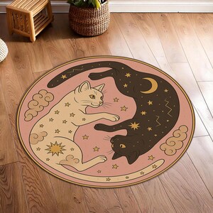 Pode incluir: Tapete redondo com fundo rosa apresentando dois gatos estilizados, um bege claro e outro castanho escuro, num design yin-yang. Os gatos são decorados com estrelas, nuvens e uma lua crescente.