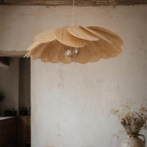 Puede incluir: Una lámpara colgante tejida con forma de flor, hecha de material natural. Tiene pétalos redondeados y superpuestos. La lámpara cuelga de un cable blanco y tiene una bombilla visible. El fondo es una pared de tonos neutros.