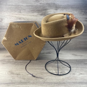 May include: A tan fedora hat with a decorative feather and bow, displayed on a black metal stand. The hat is next to a brown cardboard box with the name "MARION" and address printed in blue.
