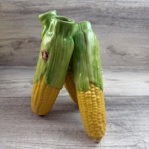 Pode incluir: Três vasos de cerâmica em forma de espiga de milho. Os vasos são pintados com grãos de milho amarelos e folhas verdes. Um pequeno adesivo é visível em um vaso. Os vasos estão dispostos sobre uma superfície de madeira cinza claro.