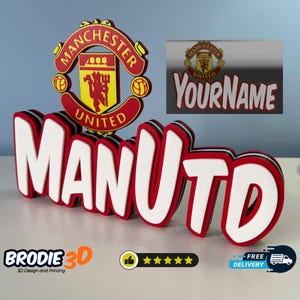 May include: A 3D printed name sign with the word "MANUTD" in red and white letters. The letters are layered and have a black outline. The Manchester United crest is in the background.