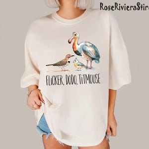 May include: A white t-shirt with a watercolor illustration of a dodo, a flicker, and a titmouse. The text "Flicker, Dodo, Titmouse" is printed below the birds.