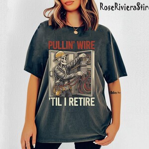 May include: A dark gray t-shirt with a distressed look features a skeleton electrician working on a fuse box. The text on the shirt reads "Pullin' Wire 'Til I Retire".
