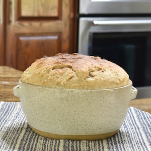 May include: A round loaf of bread in a white ceramic baking dish with brown speckled glaze. The dish has two handles on the sides. The bread is sitting on a blue and white striped kitchen towel.