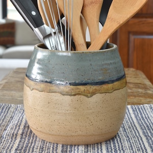 May include: A handmade ceramic utensil holder with a speckled tan base, a blue-gray top, and a dark blue band. The holder contains various kitchen tools, including a whisk, spatulas, and knives. The holder sits on a striped blue and white cloth.