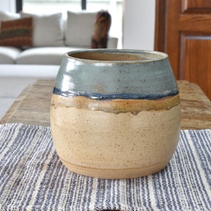 May include: A handcrafted ceramic vase with a wide, rounded shape. The top is glazed in a light blue, transitioning to a tan speckled body. A thin blue band separates the colors. The vase sits on a striped blue and white table runner.