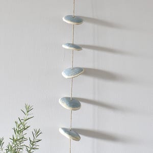 May include: A vertical hanging decoration featuring six smooth, circular ceramic discs in a pale blue glaze, each with a white edge. The discs are evenly spaced and strung together on a thin, light brown natural fiber cord, creating a linear display.
