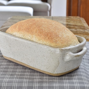 May include: A loaf of freshly baked bread in a speckled ceramic bread pan. The bread is golden brown, and the pan has handles on each side. The pan is sitting on a gray and white plaid cloth.