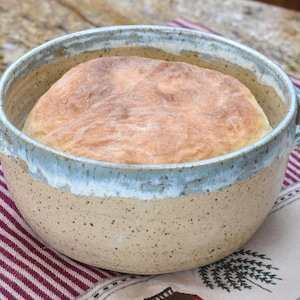 May include: A round, light brown ceramic bread baking dish with a light blue glaze around the rim. The dish has two handles and is filled with a freshly baked loaf of bread.