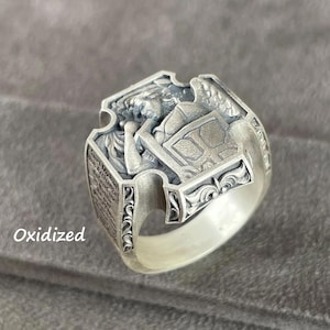 May include: Oxidized silver ring with a detailed, embossed design. The ring features a central rectangular emblem with a figure and text, surrounded by ornate scrollwork and a textured band. The ring is set against a gray background.