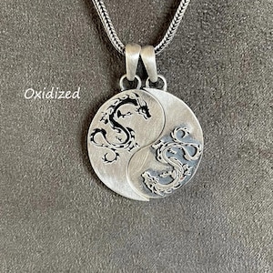 May include: A silver pendant necklace with a Yin and Yang design, featuring dragon illustrations. The round pendant has a textured finish and a silver chain. The word "Oxidized" is printed on the left side of the image.