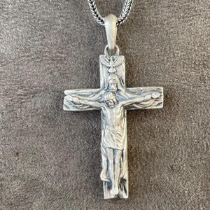 May include: A silver-colored pendant necklace featuring a detailed crucifix design. The pendant depicts a figure on the cross, with intricate detailing. The necklace chain is a woven style, and the background is a textured gray surface.